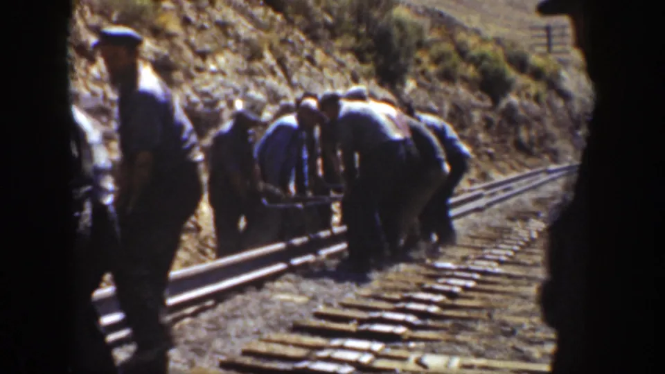 A worker stands near the camera as others bend over rails and ties on a dry 1936 California railroad work site.