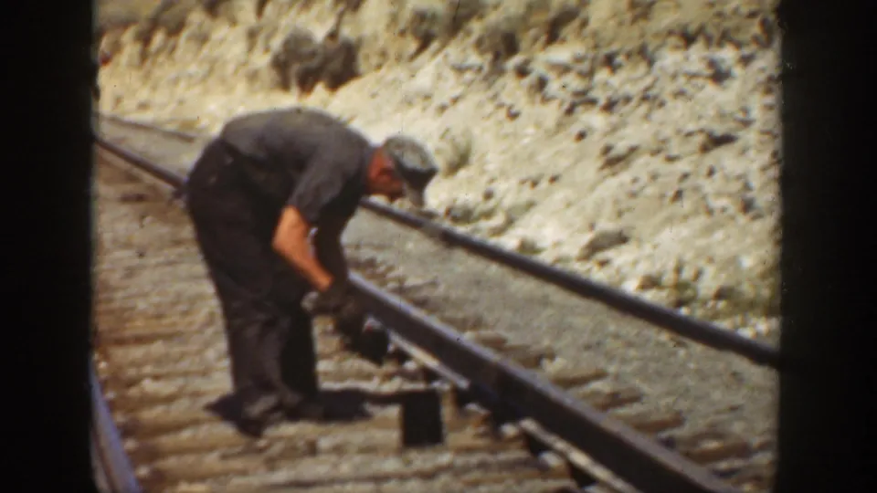 In 1936 California footage, a railroad worker in dark clothes bends over the rail while working near wooden ties and dry earth.