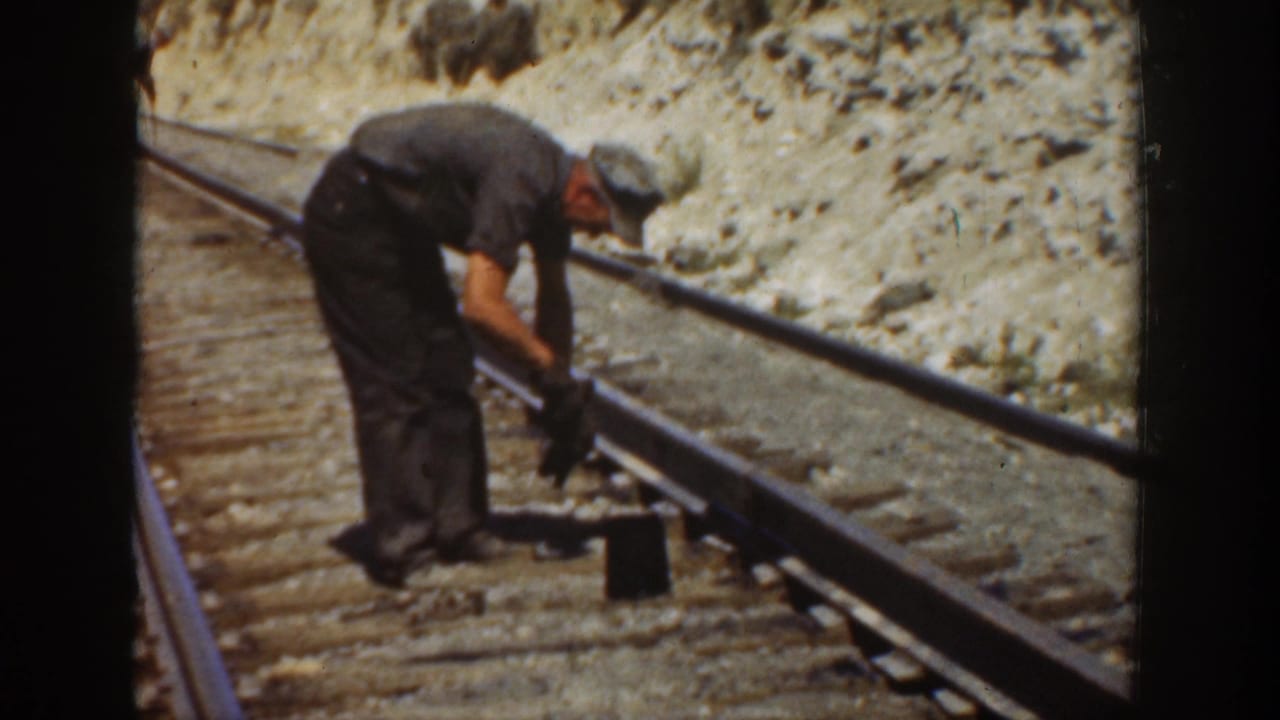 A railroad worker appears to adjust or inspect a rail with a hand tool on a sunlit California track bed.