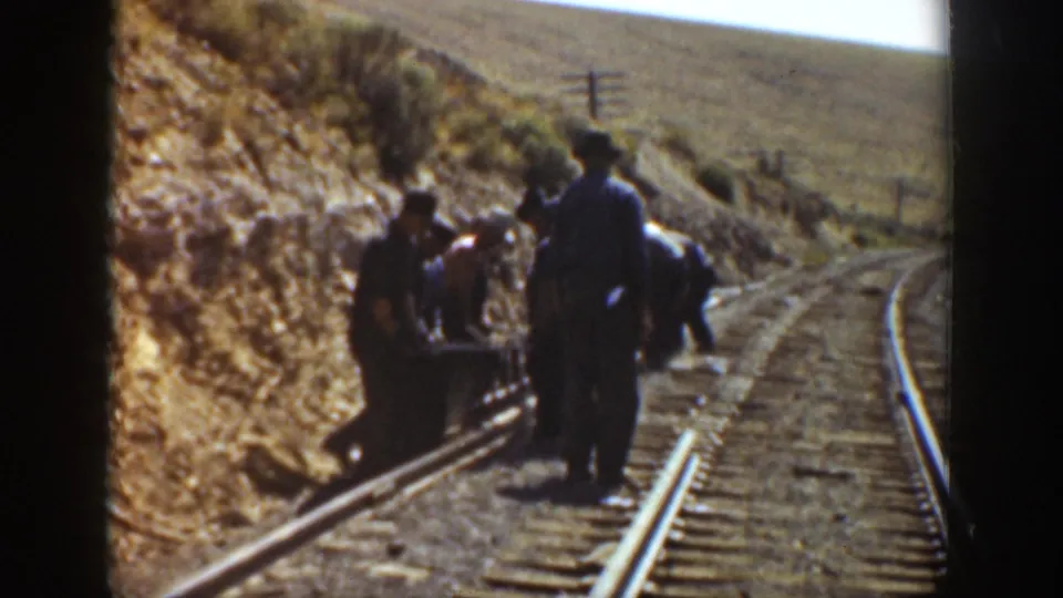 Several workers stand and bend over the rails as the track curves through a sunlit, arid California landscape.