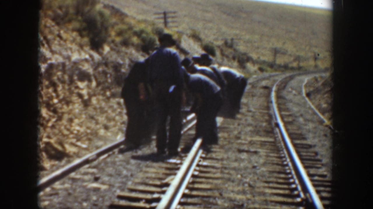 Several railway workers in dark clothing work beside curved tracks in rural California, with dry hills and utility poles visible.