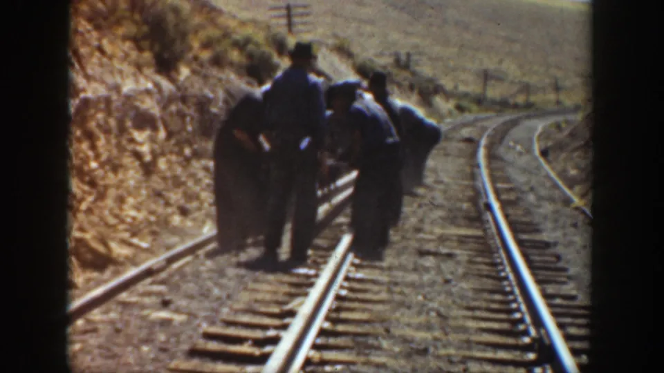 A railway worker stands near others bending over the track, with rails, ties, and dry California hillside visible in 1936 footage.