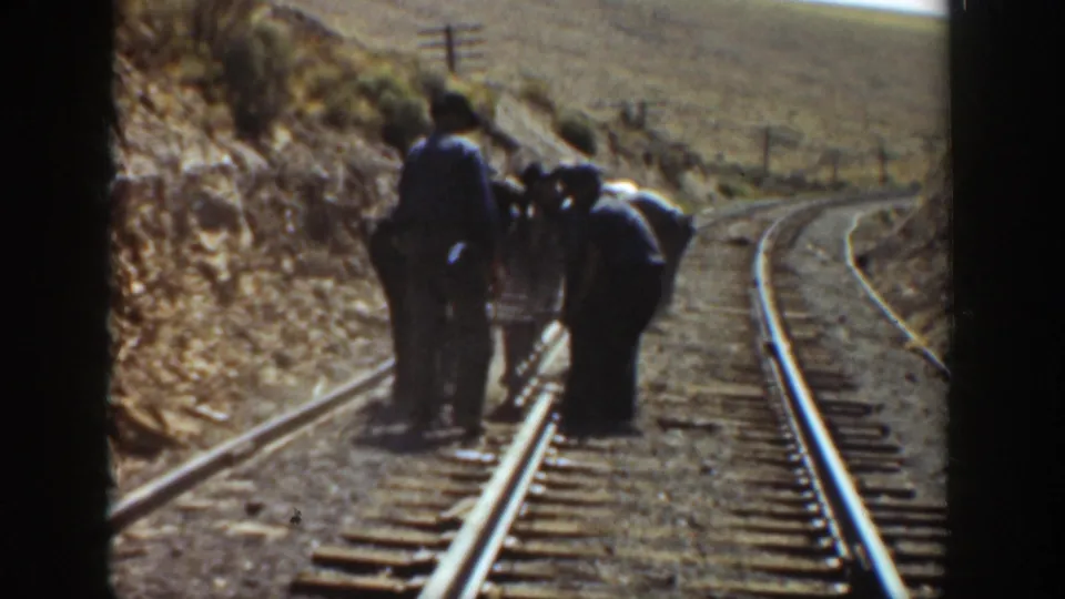 Railway workers continue track maintenance on a curved California line, framed by dry hills and the visible edge of 8mm film.