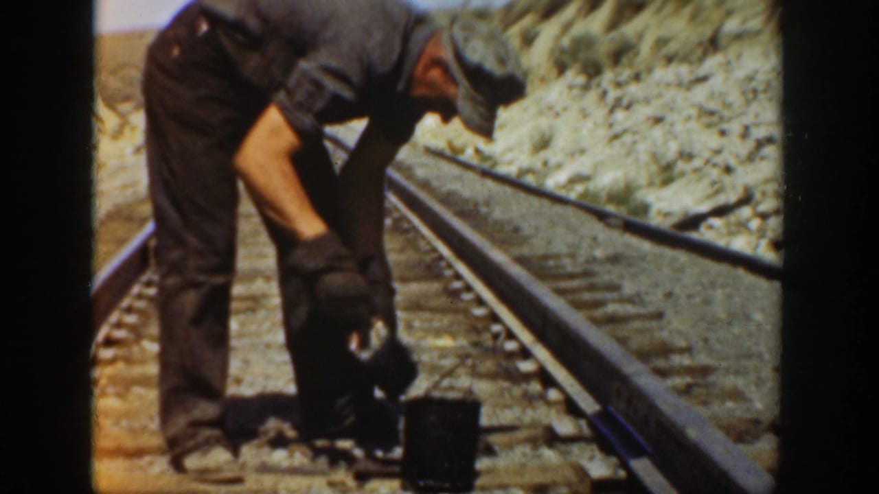 1936 California 8mm frame showing a gloved man bent over beside railroad rails in a dry outdoor setting.