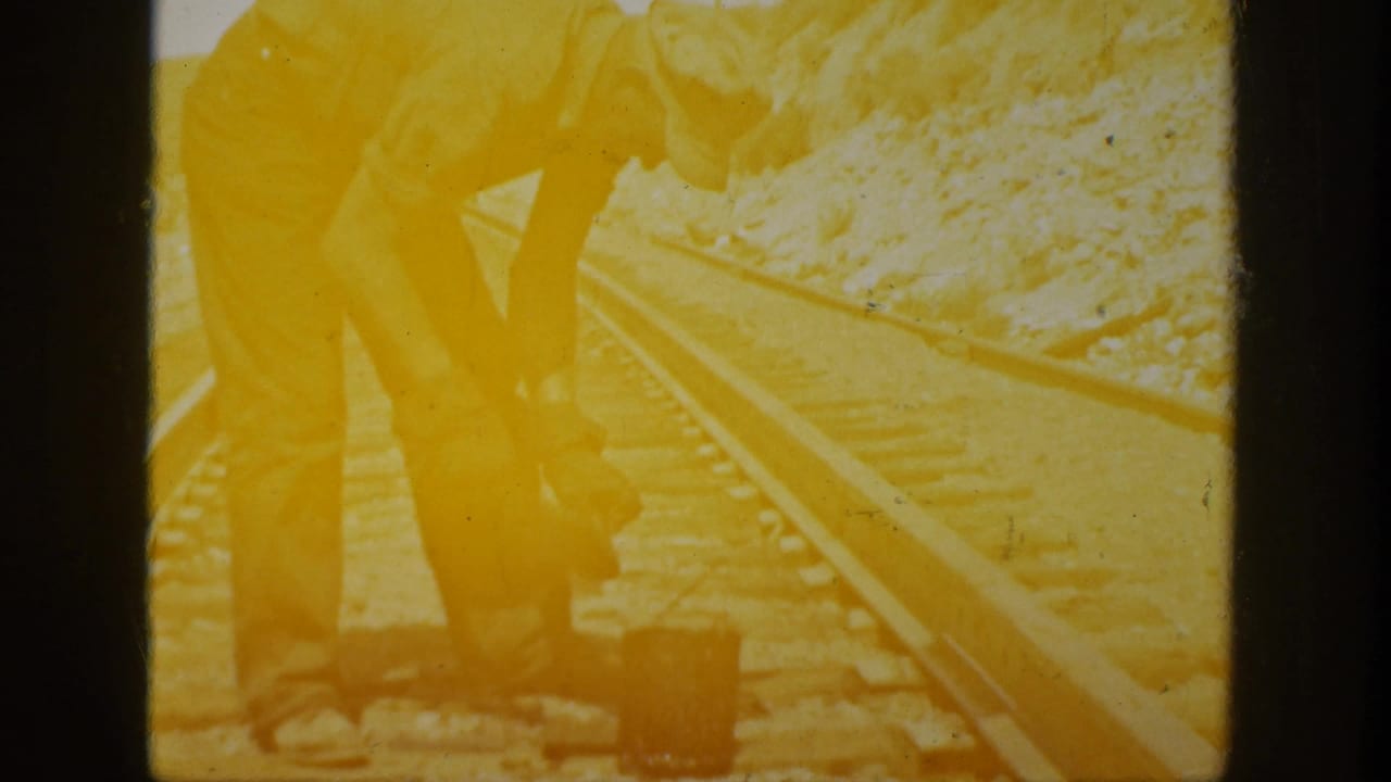 Warm, overexposed 8mm frame of the track worker beside rails, with arid ground and strong sunlight visible.