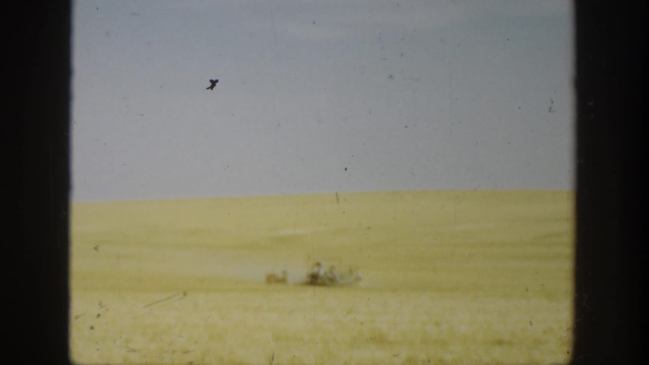 1936 California home-movie still with a pale wheat field, overcast sky, and tiny indistinct people or machinery far away.