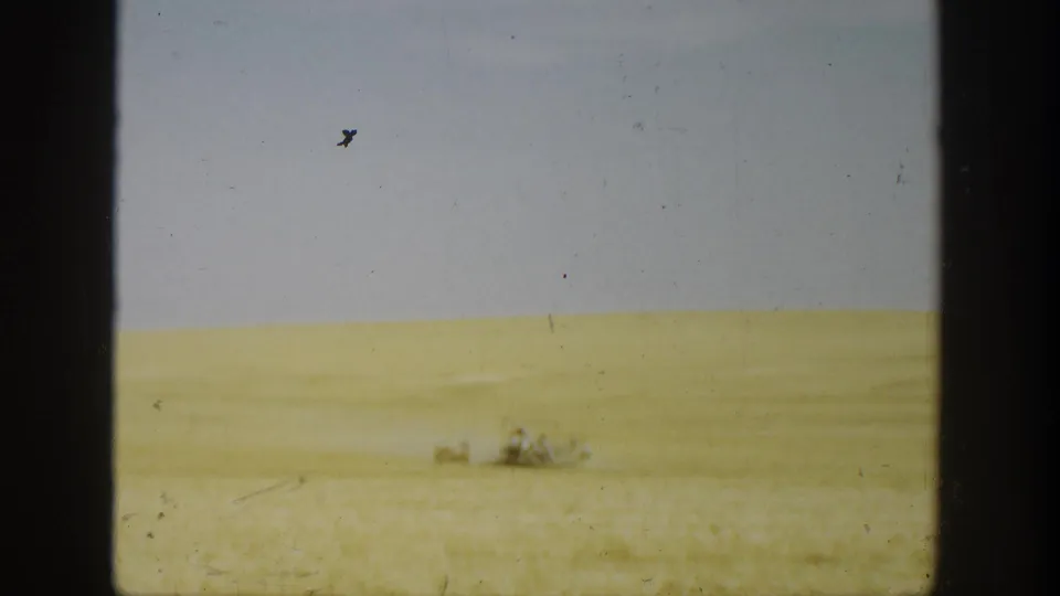 1936 California home-movie still with a pale wheat field, overcast sky, and tiny indistinct people or machinery far away.