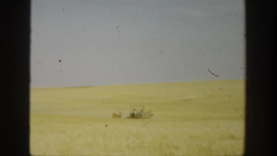 1936 California home-movie frame of an open wheat field with tiny distant figures or equipment and visible 8mm film wear.