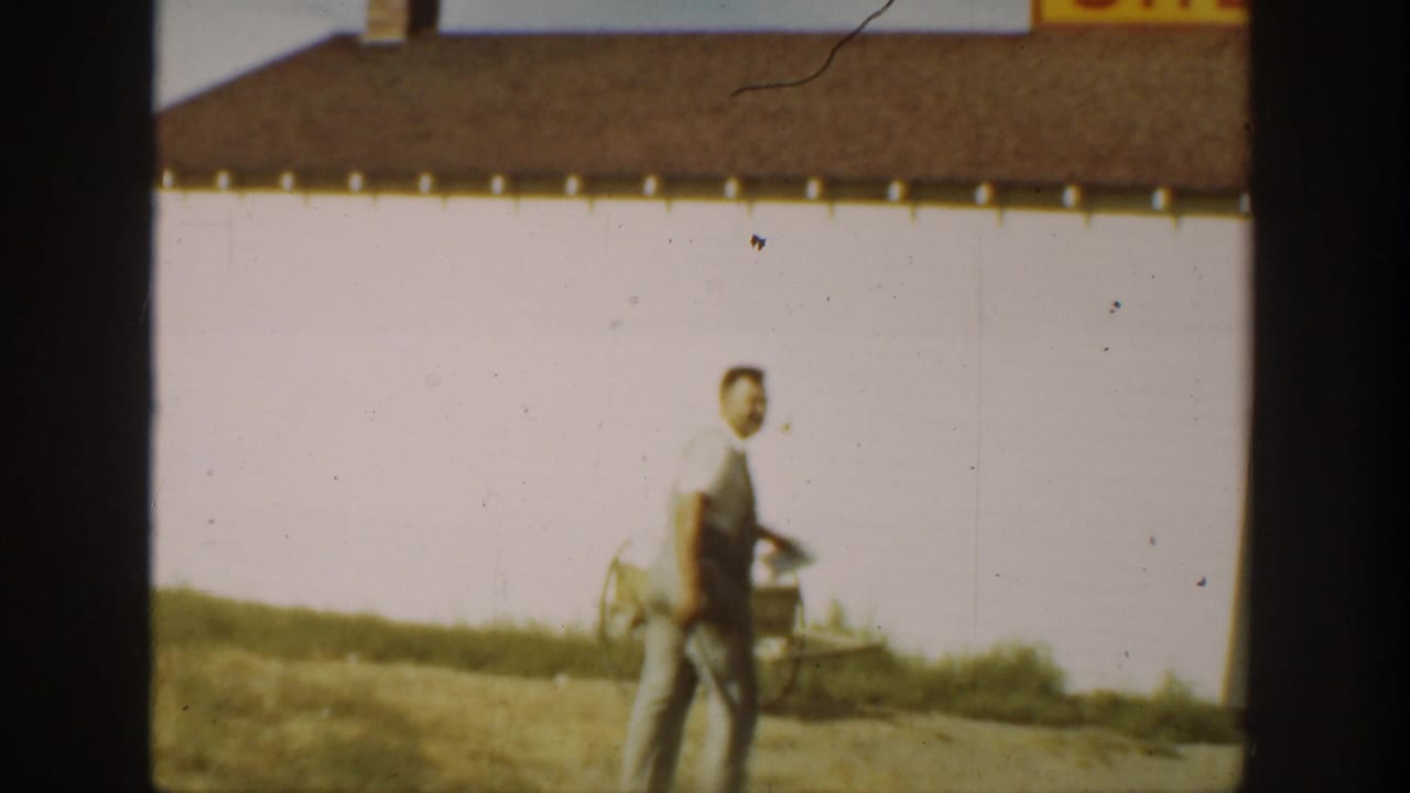 Adult male farmer in overalls walks near a white barn in 1930s California home-movie footage, carrying a hand tool at his side.