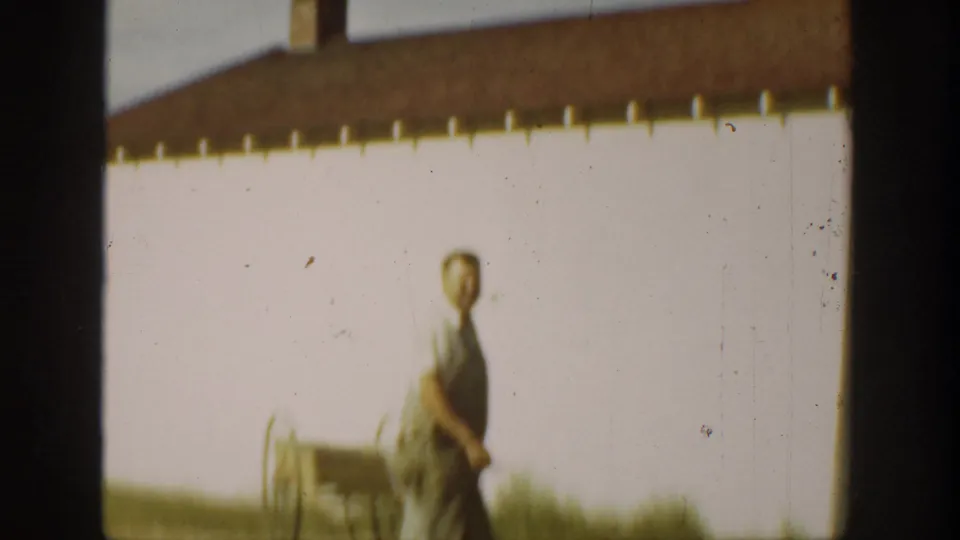 Adult farmer in overalls pauses or walks near a white barn, with the soft focus and color fading of 1930s California 8mm footage.