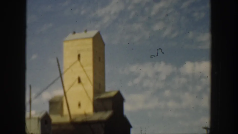 Wider 1936 California frame showing a tall ore-processing or industrial tower with adjoining low buildings and open dirt ground.