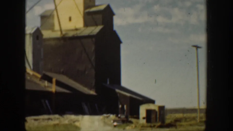 1936 California home-movie frame showing a small industrial or mine-processing complex with tower, sheds, open ground, and utility pole.