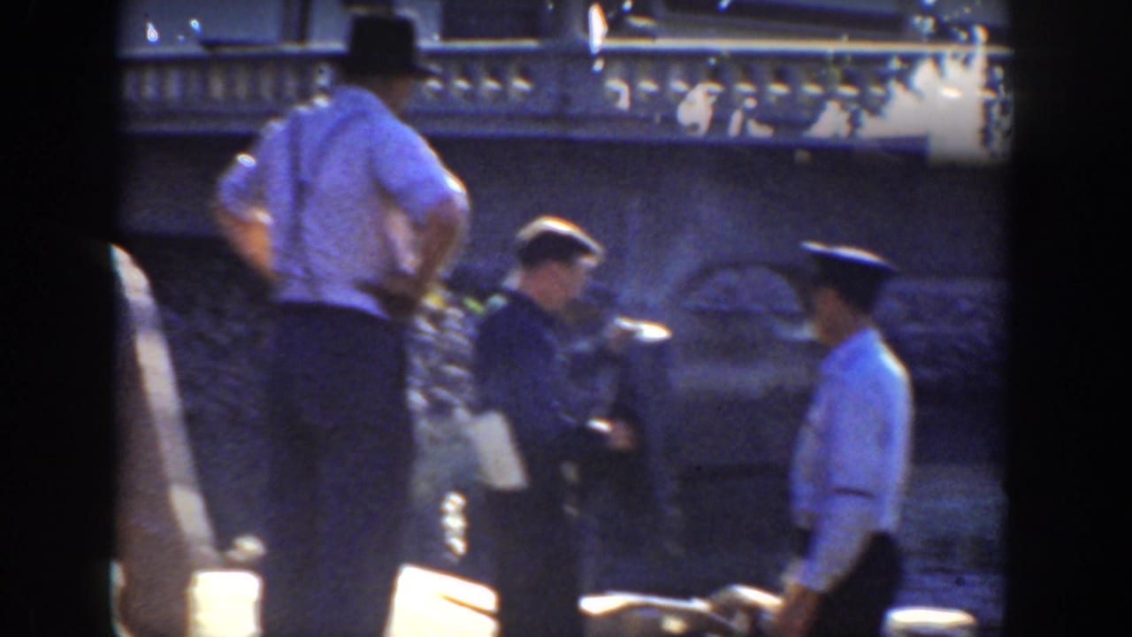 1936 California home-movie frame showing men in period hats near a river or dock, with a stone bridge or embankment in the background.