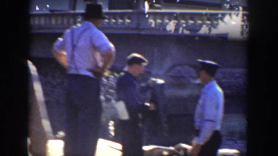 1936 California home-movie frame showing men in period hats near a river or dock, with a stone bridge or embankment in the background.