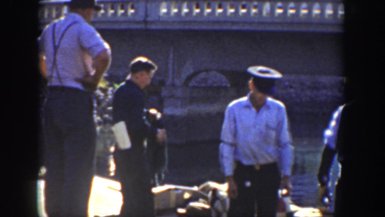 1936 California frame with men in period clothing beside rippled water and a stone bridge or embankment, including a police-style uniformed figure.