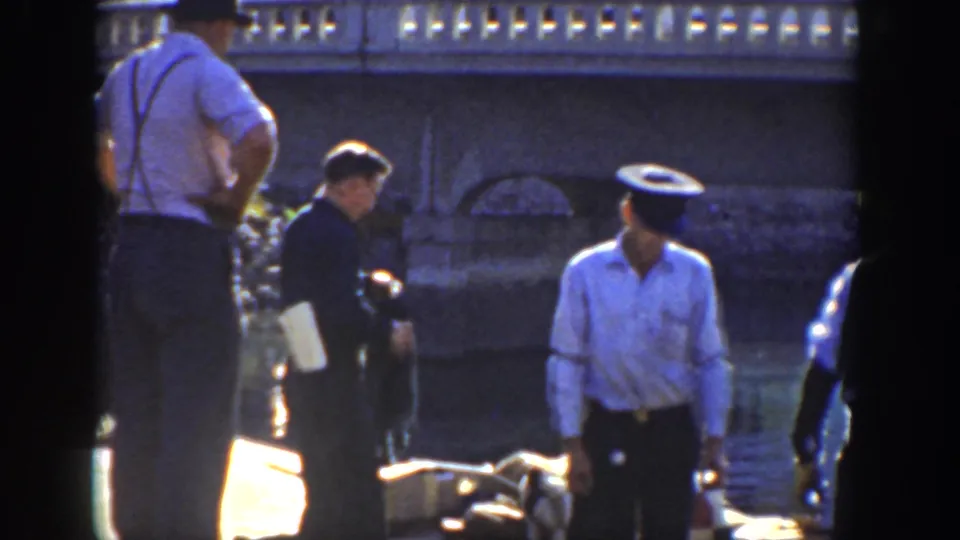 1936 California frame with men in period clothing beside rippled water and a stone bridge or embankment, including a police-style uniformed figure.