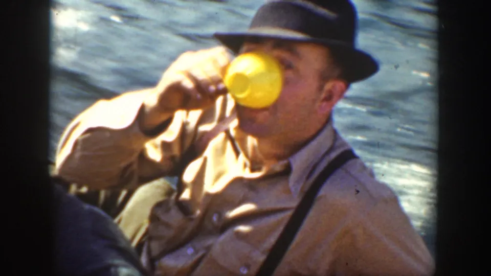 1936 California home-movie frame of a seated man in a fedora and suspenders drinking from a yellow cup on a boat over rippled water.