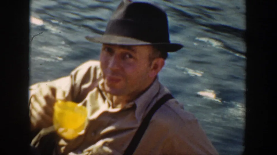 1936 California frame of a man in a fedora and suspenders holding a yellow cup on a boat, with sunlit water behind him.