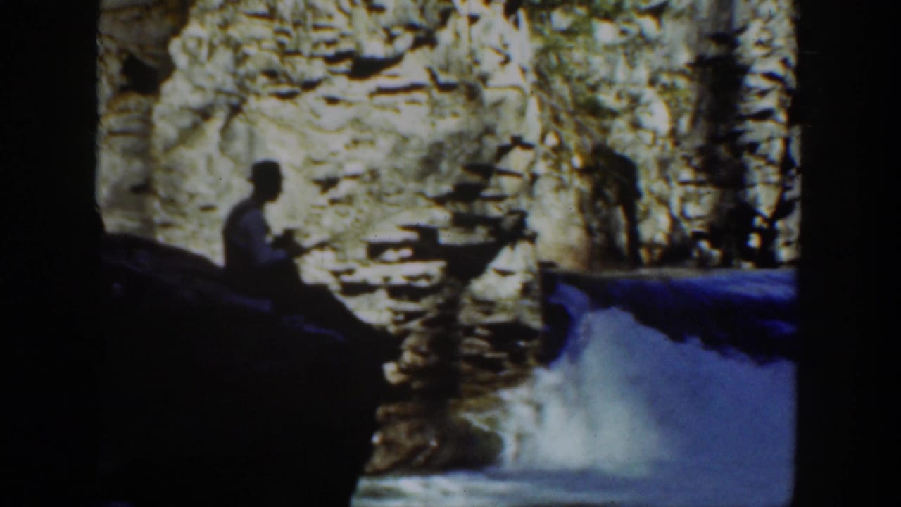 1936 California home-movie frame with a man seated near a creek, silhouetted against rocks and fast-moving water.