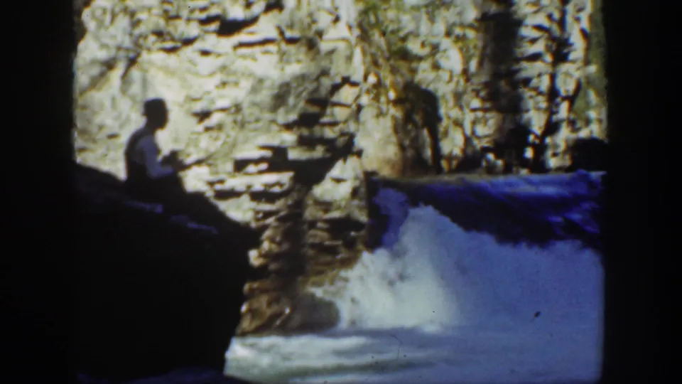 1936 California 8mm frame showing a seated angler beside bright rushing creek water and layered rock formations.