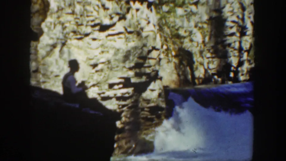 1936 California home-movie frame of a man near a creek, with blue-toned water rushing past a stone and rock backdrop.