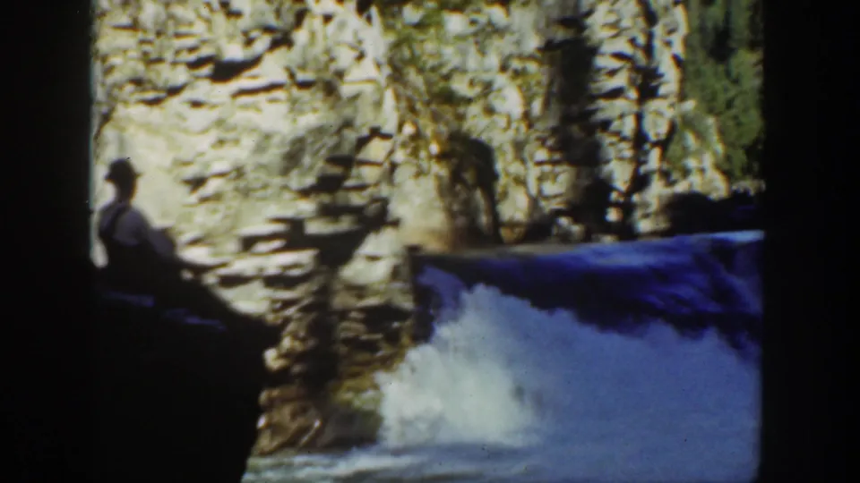 A 1936 California home-movie frame places the seated visitor at left against a bright waterfall and rugged stone wall.