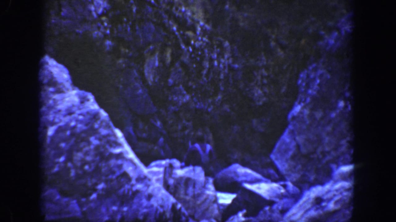Blue-tinted 8mm frame showing rocky formations and boulders, possibly water or cave imagery, before the canyon scene changes.