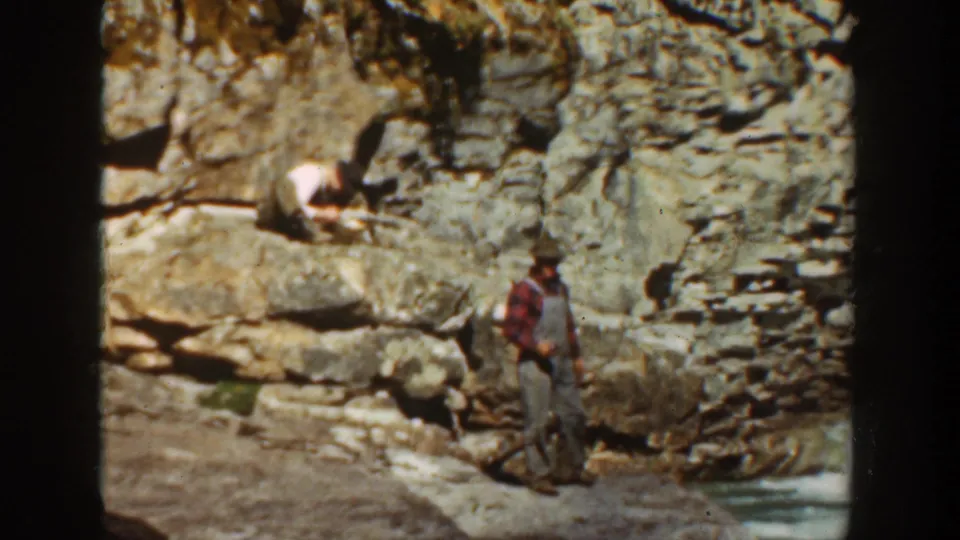 1936 California 8mm frame showing two adult men at a rocky canyon water area, with one standing in plaid and another crouched on the rocks.