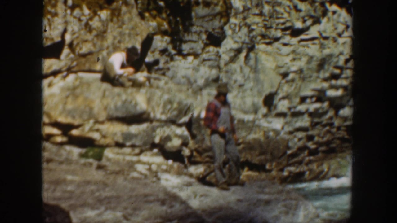 1936 California home-movie frame showing two men by layered coastal rocks, with one standing below a seated figure on a sunny cliffside.