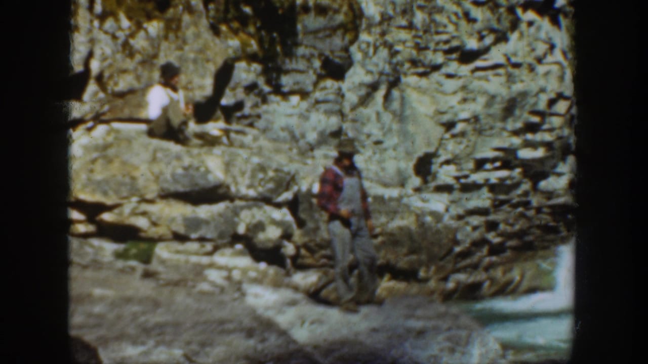 A man in plaid and overalls stands near a rocky cliff while another person sits higher on the ledge in 1936 California footage.