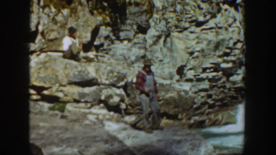 A man in plaid and overalls stands near a rocky cliff while another person sits higher on the ledge in 1936 California footage.