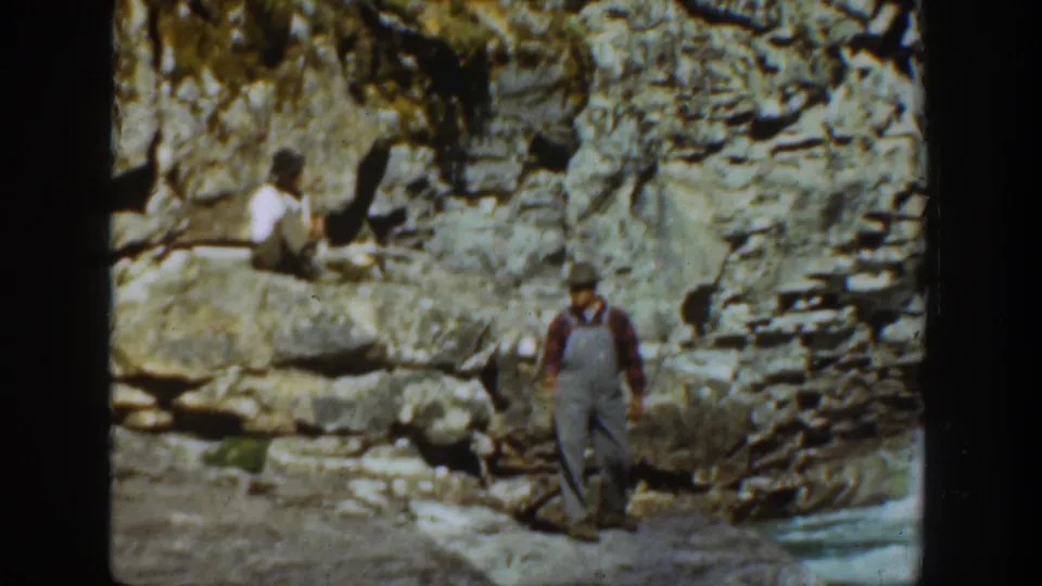 The standing man faces toward the camera near cliffside stones as a seated person remains on the ledge above in the California home movie.