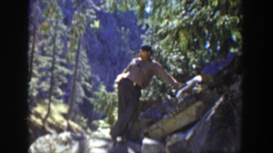 1936 California frame of a man in a light shirt and dark trousers balancing on boulders near a mountain stream.