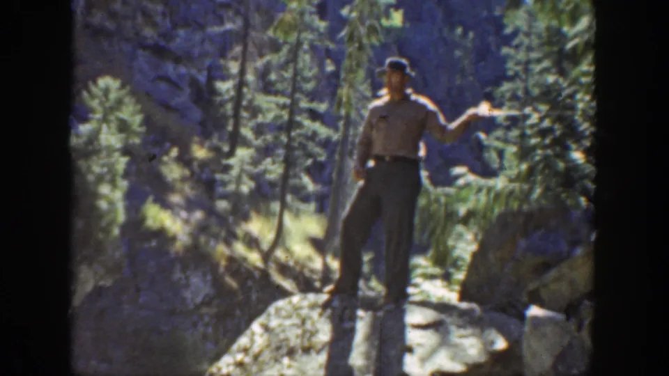 A man in a hat and light shirt stands on a boulder with a fishing rod extended in 1930s California stream footage.