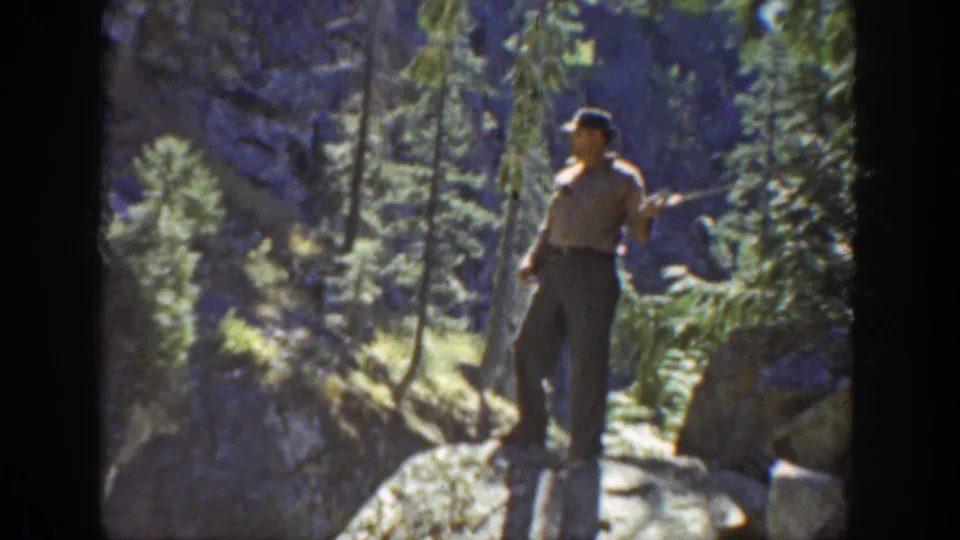 The fisherman stands on rocks beside a rushing mountain stream, framed by conifers and rugged terrain in 1936 California footage.