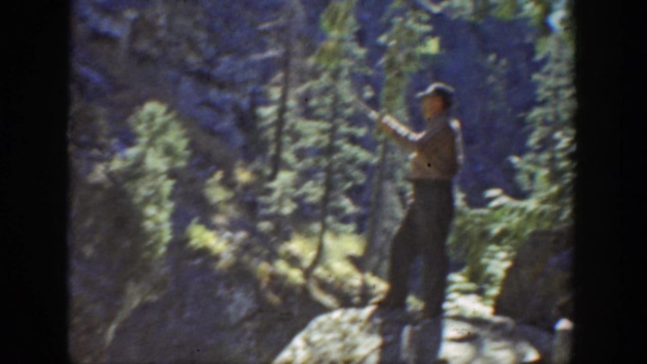 1936 California home-movie frame of a man with a long fishing rod standing among rocks and evergreen trees.