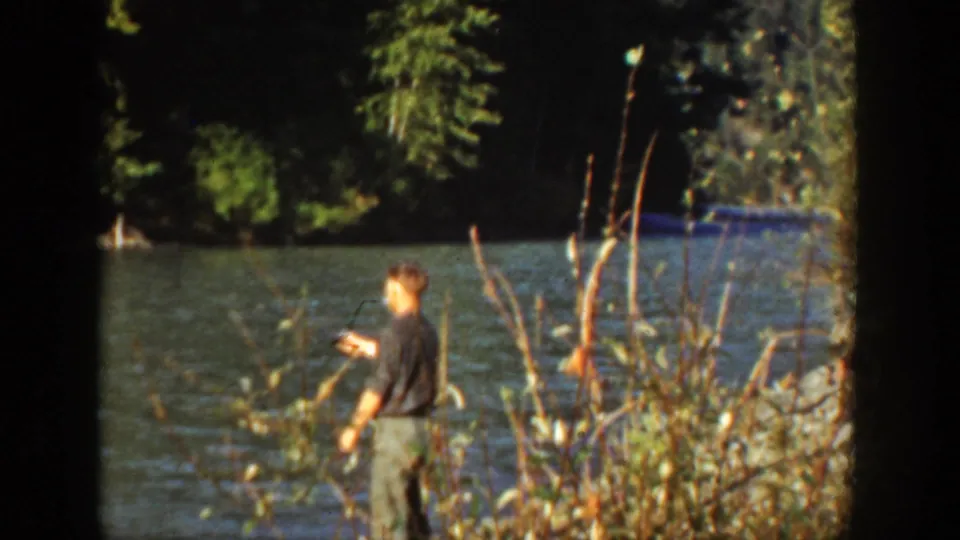 1936 California lakeside frame showing a man fishing near brush with trees and water behind him.
