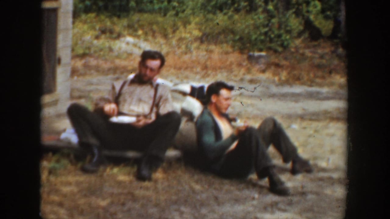 1936 California 8mm frame of two men resting outside, with one seated by a low bench eating from a plate and another leaning back on dry ground.