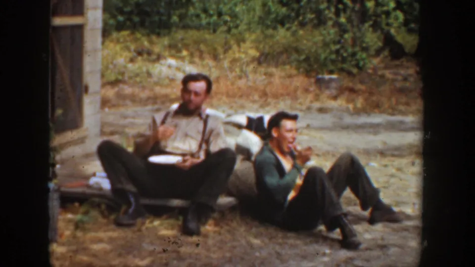 1936 California 8mm frame showing two men seated outside, one eating from a plate while the other rests beside him near a small structure.