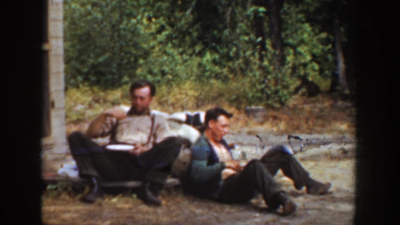 Two men sit on the ground in a wooded area, with one holding a plate and the other leaning back.
