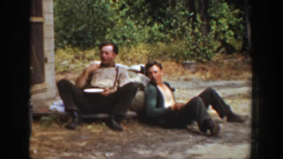 California 1936 home-movie frame of two adult men pausing outdoors, one eating and one lounging on the dry ground.