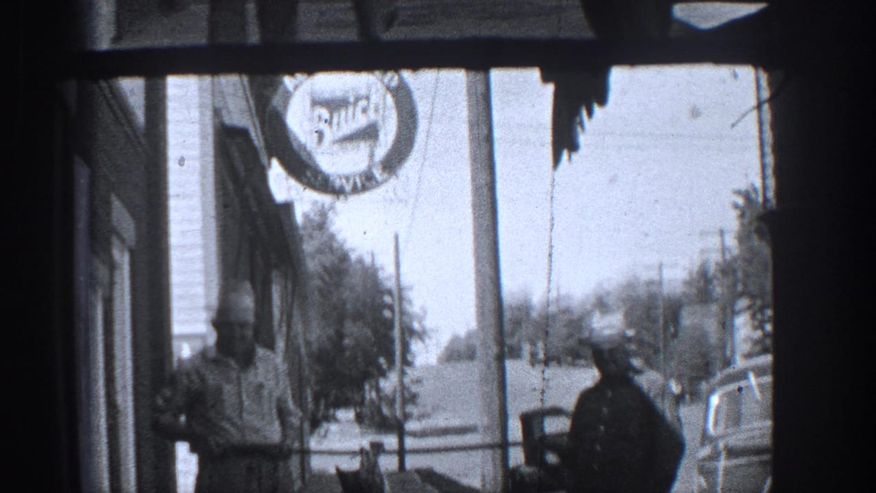 Two men stand near a storefront with a circular sign, sidewalk, cars, and utility poles visible.
