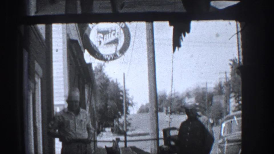 1936 California street view with two men near a storefront, a circular sign, parked cars, and utility poles.