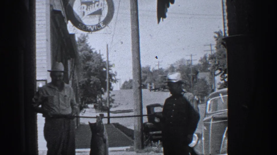 1936 California home-movie frame showing a large fish held on a pole beside a uniformed man on a town sidewalk.