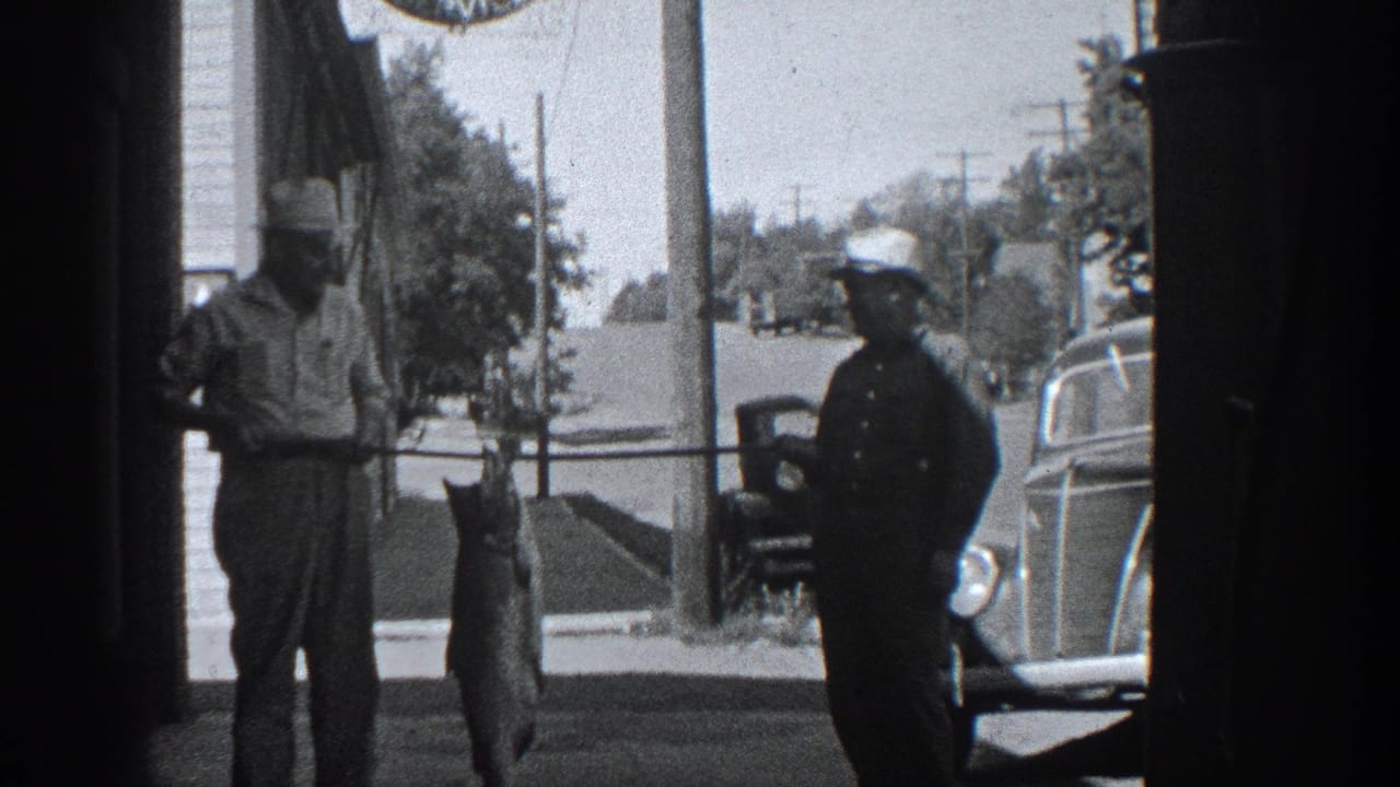 In 1936 California home-movie footage, two men pose on a sidewalk with a large fish suspended from a pole near parked cars.