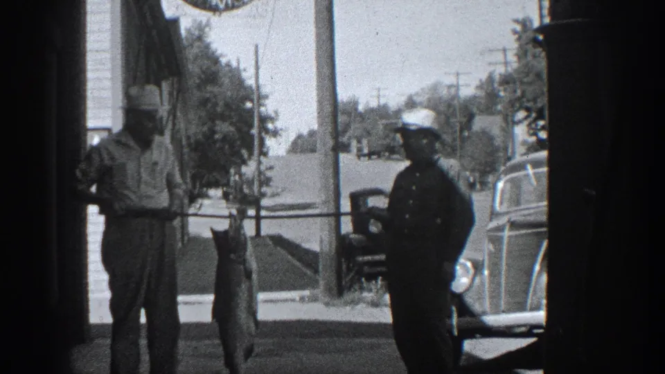 In 1936 California home-movie footage, two men pose on a sidewalk with a large fish suspended from a pole near parked cars.