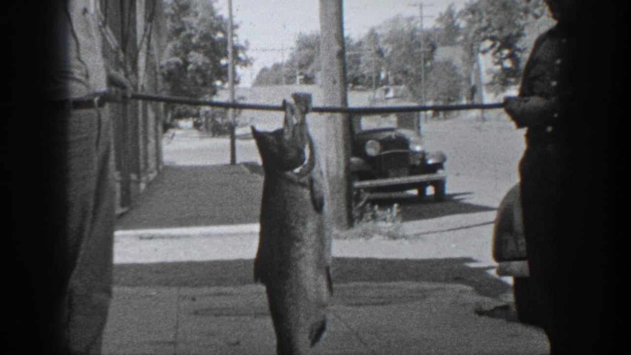 A large fish hangs from a pole beside a man, with a parked car in the street.