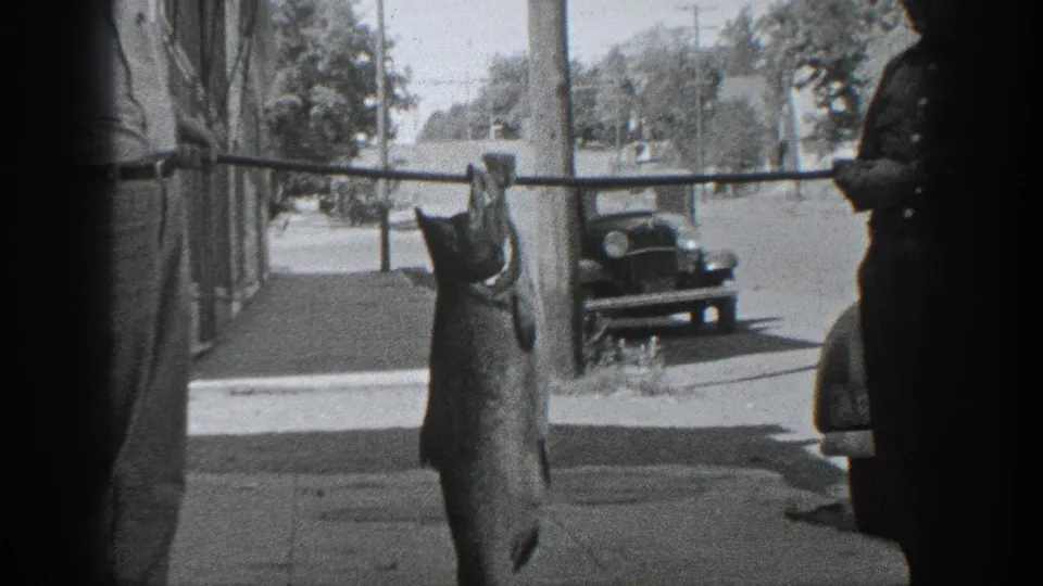 A large fish hangs prominently from a pole in a 1930s California street scene, with a man and vintage automobile nearby.