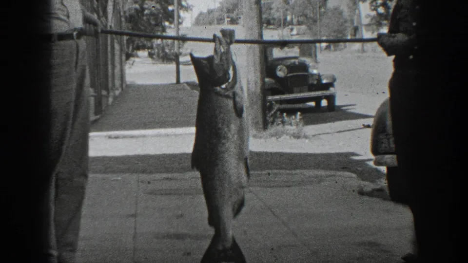 The home-movie frame centers the large hanging fish on a sidewalk, with a man partly visible and a vintage car across the curb.