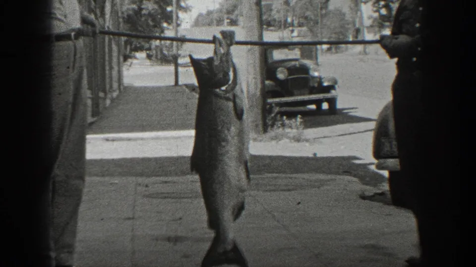 A 1936 California home-movie frame shows a large fish suspended from a pole on a sidewalk, with a man and parked automobile nearby.
